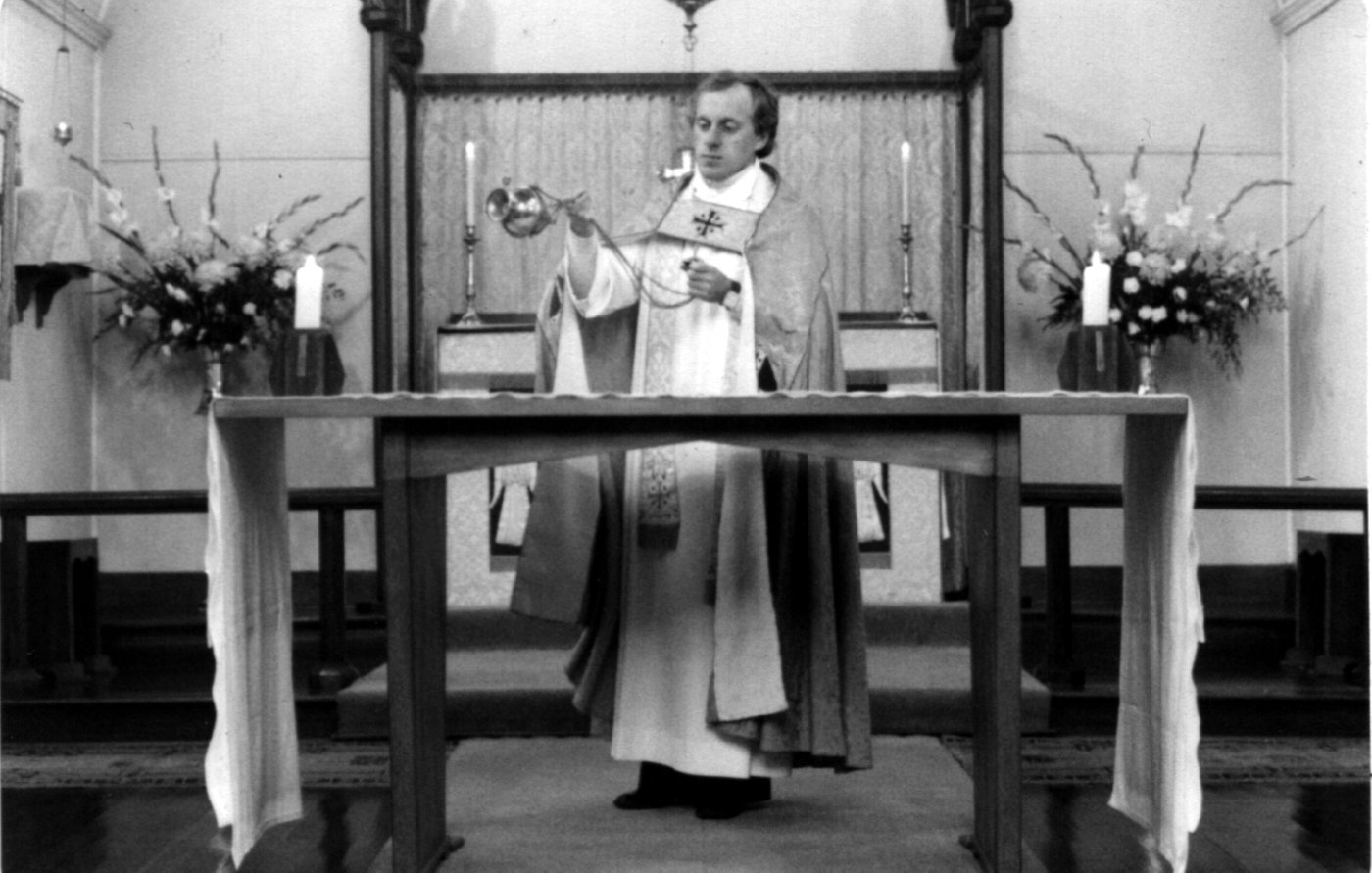 1977 Ron Dowling
In October, 1975, Bishop Grant (the Regional Bishop) proposed a young priest, the Reverend Ron Dowling who would like to come to St Linus for about 12 months.  Ron's appointment as PIC in February 1976 created an air of expectation; the vicarage had been brought up to date and a young priest in his first parish had chosen St Linus’, albeit on a third-time basis.  Ron was an ideal choice.  He was musical, had a good understanding of Liturgy and was hard working and responsible.  After years of relative seclusion the parish was exposed to the wider diocese. Part of Ron's vision was to bring to the parish up to date ideas of worship.  He revamped the order of service and worked to create a worship space which suited modern liturgy. In 1978, following the success of a planned giving program, the church building was finally completed. When Ron Dowling left the parish (now 1/2 time) on Christmas Day, 1978 to pursue his MA overseas, he left a parish that was markedly different to the one he joined in 1975.
This photo, taken in 1977, predates the church building completion and shows Ron leading the service at the old altar at the eastern end of the church.
